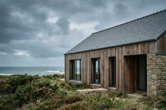 Maison passive moderne en Bretagne avec bardage bois et toiture en ardoise, entourée de végétation côtière sous un ciel nuageux atlantique