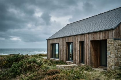 Maison passive moderne en Bretagne avec bardage bois et toiture en ardoise, entourée de végétation côtière sous un ciel nuageux atlantique